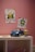 A child's room with pink walls featuring two colorful drawings of a butterfly and a flower on the wall. A small blue toy convertible car and a toy character are placed on a white shelf.