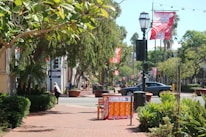 A sunny street scene featuring lush green trees along the sidewalk, large potted plants, and red patterned banners hanging from street lamps. A person is walking near a building with 'For Lease' signage. Cars are on the road, and there are people in the distance.