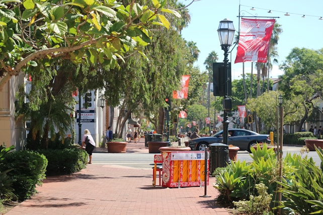 A sunny street scene featuring lush green trees along the sidewalk, large potted plants, and red patterned banners hanging from street lamps. A person is walking near a building with 'For Lease' signage. Cars are on the road, and there are people in the distance.