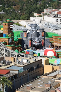 A theme park area resembling a video game landscape with various elements like a castle, colorful blocks, and large mushrooms. The setting includes animated characters and props designed to create an immersive atmosphere. Buildings and structures, including pipes and towers, are visible, surrounded by lush greenery and city infrastructure.