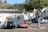 A parking lot with two cars parked next to a small building. The left car is black and the right car has a unique design with a pizza-themed wrap. There are several signs on the wall of the building indicating parking regulations. A tree is situated nearby, casting a shadow on the ground. Residential buildings are visible in the background, along with a hill covered in trees.
