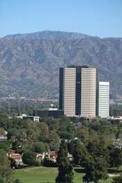 A tall office building complex stands prominently in the middle of a cityscape, surrounded by lush green trees and smaller residential buildings. In the background, a range of mountains stretches across the horizon under a clear blue sky, providing a scenic backdrop.