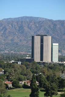 A tall office building complex stands prominently in the middle of a cityscape, surrounded by lush green trees and smaller residential buildings. In the background, a range of mountains stretches across the horizon under a clear blue sky, providing a scenic backdrop.