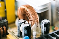 Close-up of electronic components on a circuit board, featuring a large copper coil and several capacitors. The composition reveals intricate details and textures of the metal and plastic parts.