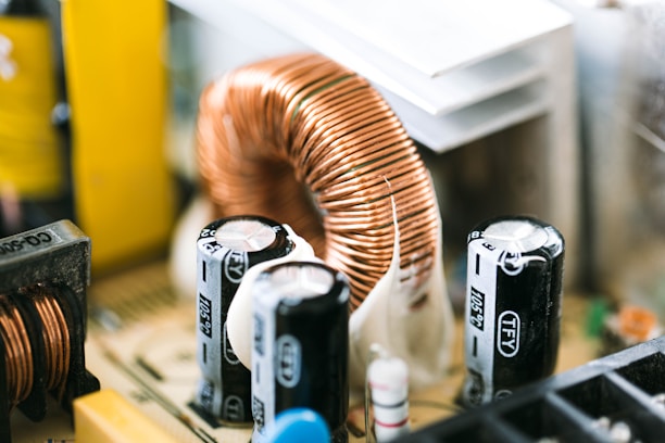 Close-up of electronic components on a circuit board, featuring a large copper coil and several capacitors. The composition reveals intricate details and textures of the metal and plastic parts.