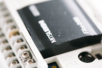 A close-up view of an industrial electronic component labeled with the brand Mitsubishi. The component is mounted in a white casing with several screws and connectors visible.