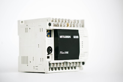 A gray industrial electronic device with multiple ports on the top and a black panel on the front labeled 'Mitsubishi' and 'FX3G-24M'. The unit has a rectangular shape with various connectors exposed, indicating its use for automation or control purposes.