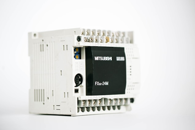 A gray industrial electronic device with multiple ports on the top and a black panel on the front labeled 'Mitsubishi' and 'FX3G-24M'. The unit has a rectangular shape with various connectors exposed, indicating its use for automation or control purposes.