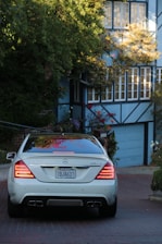 A silver Mercedes-Benz S63 AMG is parked on a red-brick driveway, surrounded by greenery and adjacent to a house with a blue garage door and numerous windows. The environment appears residential, with lush plants and a tree partially covering the building.