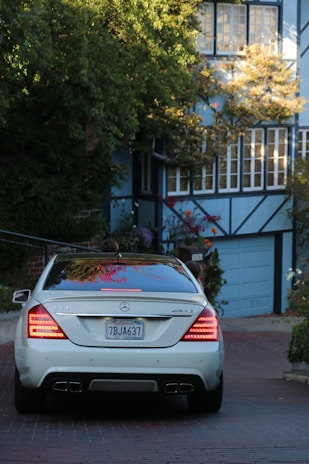 A silver Mercedes-Benz S63 AMG is parked on a red-brick driveway, surrounded by greenery and adjacent to a house with a blue garage door and numerous windows. The environment appears residential, with lush plants and a tree partially covering the building.