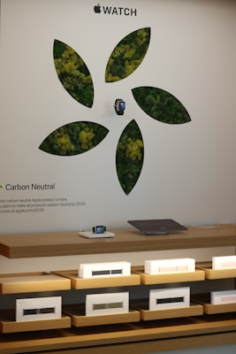 Promotional display featuring an Apple Watch with a design that includes green and yellow foliage motifs in a pattern resembling leaves. Below the watch is a shelf with more Apple Watches and accessories, set against a white wall that highlights a message about carbon neutrality.