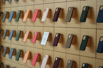 A wooden wall displays an organized array of phone cases in various colors, arranged on square panels. Each case is mounted vertically, showcasing a spectrum of hues including green, orange, pink, and blue. A small sign labeled 'Self-Checkout Available' is positioned among the cases.