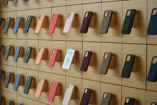 A wooden wall displays an organized array of phone cases in various colors, arranged on square panels. Each case is mounted vertically, showcasing a spectrum of hues including green, orange, pink, and blue. A small sign labeled 'Self-Checkout Available' is positioned among the cases.