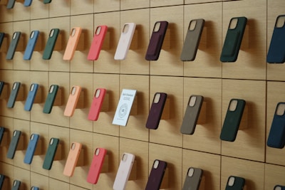 A wooden wall displays an organized array of phone cases in various colors, arranged on square panels. Each case is mounted vertically, showcasing a spectrum of hues including green, orange, pink, and blue. A small sign labeled 'Self-Checkout Available' is positioned among the cases.