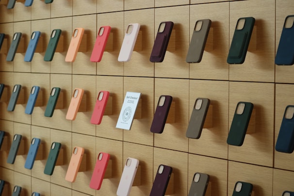 A wooden wall displays an organized array of phone cases in various colors, arranged on square panels. Each case is mounted vertically, showcasing a spectrum of hues including green, orange, pink, and blue. A small sign labeled 'Self-Checkout Available' is positioned among the cases.