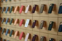A wooden wall displays an organized array of phone cases in various colors, arranged on square panels. Each case is mounted vertically, showcasing a spectrum of hues including green, orange, pink, and blue. A small sign labeled 'Self-Checkout Available' is positioned among the cases.