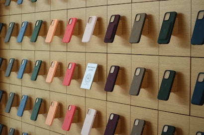 A wooden wall displays an organized array of phone cases in various colors, arranged on square panels. Each case is mounted vertically, showcasing a spectrum of hues including green, orange, pink, and blue. A small sign labeled 'Self-Checkout Available' is positioned among the cases.