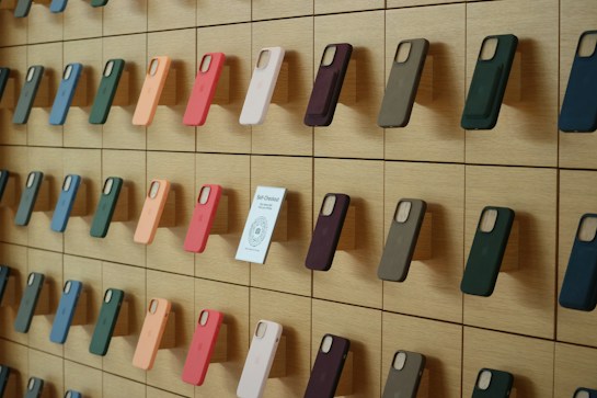 A wooden wall displays an organized array of phone cases in various colors, arranged on square panels. Each case is mounted vertically, showcasing a spectrum of hues including green, orange, pink, and blue. A small sign labeled 'Self-Checkout Available' is positioned among the cases.