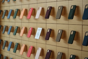 A wooden wall displays an organized array of phone cases in various colors, arranged on square panels. Each case is mounted vertically, showcasing a spectrum of hues including green, orange, pink, and blue. A small sign labeled 'Self-Checkout Available' is positioned among the cases.