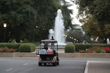 A golf cart carries two people toward a large, elegantly arched fountain, surrounded by manicured hedges and vibrant flower beds. The pathway and nearby streetlamps create a serene ambiance under a canopy of lush green trees.