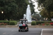A golf cart carries two people toward a large, elegantly arched fountain, surrounded by manicured hedges and vibrant flower beds. The pathway and nearby streetlamps create a serene ambiance under a canopy of lush green trees.