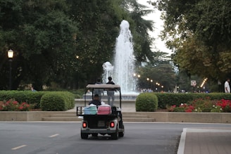 A golf cart carries two people toward a large, elegantly arched fountain, surrounded by manicured hedges and vibrant flower beds. The pathway and nearby streetlamps create a serene ambiance under a canopy of lush green trees.