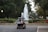 A golf cart carries two people toward a large, elegantly arched fountain, surrounded by manicured hedges and vibrant flower beds. The pathway and nearby streetlamps create a serene ambiance under a canopy of lush green trees.