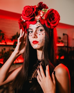 A woman with long dark hair is adorned with a crown of large red roses and smaller flowers. Her face is intricately painted with white and red decorative patterns, resembling traditional festive makeup. She wears a black top and has dark nail polish, standing against a background lit with warm red lights.