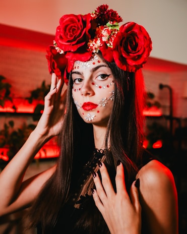 A woman with long dark hair is adorned with a crown of large red roses and smaller flowers. Her face is intricately painted with white and red decorative patterns, resembling traditional festive makeup. She wears a black top and has dark nail polish, standing against a background lit with warm red lights.