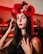 A woman with long dark hair is adorned with a crown of large red roses and smaller flowers. Her face is intricately painted with white and red decorative patterns, resembling traditional festive makeup. She wears a black top and has dark nail polish, standing against a background lit with warm red lights.