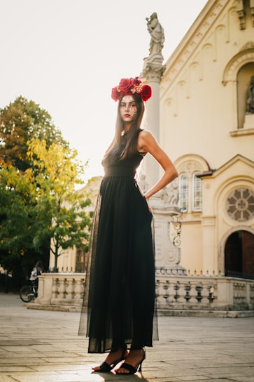 A woman stands confidently in a long black dress with floral and face-painted decoration. She wears a crown of large red roses on her head and is positioned outside in a sunlit urban setting with a historical building in the background. The lighting casts a warm glow, highlighting the intricate details of her attire.