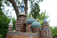A detailed close-up of a miniature castle made from clay or ceramic materials, featuring ornate spires and domes. The sculpture is decorated with intricate patterns and textures, predominantly in earthy brown, blue, and teal tones. The background consists of lush green foliage and trees.