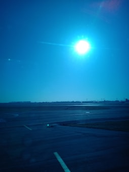 A bright sun shines in a clear blue sky above an airport runway. The tarmac is expansive, with markings visible on the surface. The horizon shows a line of distant structures, possibly airport buildings or control towers.