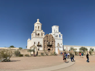 A sunlit Spanish mission framed by rugged desert hills under a vibrant blue sky.