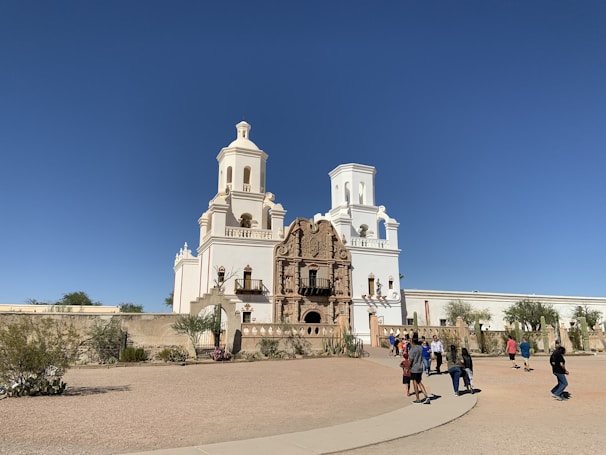 A historic mission church with white towers and ornate brown facade sits under a clear blue sky. Cacti and desert vegetation are visible in the foreground, and a few visitors walk near the entrance, adding to the liveliness of the scene.