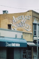 A vintage advertisement is painted on the side of a weathered brick building, featuring ornate lettering that reads 'Abstract & Guaranty Co.'. Below, storefronts with awnings display the names of various businesses, including 'Crouch Family Medicine'. The scene suggests a small-town or urban environment with a classic and historical feel.