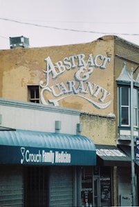 A vintage advertisement is painted on the side of a weathered brick building, featuring ornate lettering that reads 'Abstract & Guaranty Co.'. Below, storefronts with awnings display the names of various businesses, including 'Crouch Family Medicine'. The scene suggests a small-town or urban environment with a classic and historical feel.