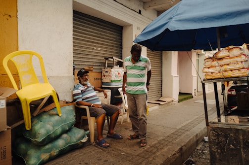 Two men are engaged in conversation near a street market setup. One man is sitting on a white plastic chair next to bags of produce and a yellow plastic chair, while the other stands nearby. A table with packaged bread is under a blue canopy. The setting is urban, with closed shop shutters and a cement walkway.