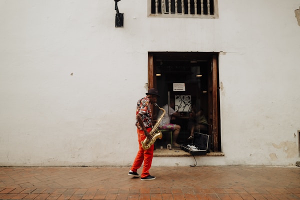 A man is standing and playing a saxophone in front of a white wall. He is wearing a floral shirt, red pants, a dark hat, and sneakers. The open window behind him reveals an interior with some furniture and a partially visible person. There is an open saxophone case on the windowsill.