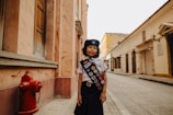 A child wearing a scout uniform with a sash adorned with colorful badges stands on a quiet street lined with old buildings. A bright red fire hydrant is visible on the left, and the architecture features warm tones and historical design elements.