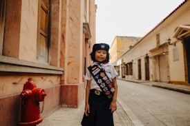 A child wearing a scout uniform with a sash adorned with colorful badges stands on a quiet street lined with old buildings. A bright red fire hydrant is visible on the left, and the architecture features warm tones and historical design elements.
