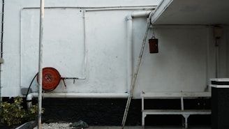 An outdoor corner with white walls and various utilitarian objects. Towards the left, there's a red hose reel attached to the wall. Pipes run along the wall horizontally and vertically. There's a hanging brown bucket, a white bench, and a sloped roof covering part of the area. To the left corner, there are green plants and gravel on the ground.