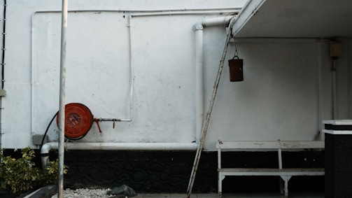 An outdoor corner with white walls and various utilitarian objects. Towards the left, there's a red hose reel attached to the wall. Pipes run along the wall horizontally and vertically. There's a hanging brown bucket, a white bench, and a sloped roof covering part of the area. To the left corner, there are green plants and gravel on the ground.