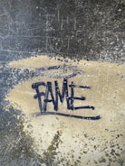 Graffiti art with the word 'FAME' spray-painted in black on a rough, textured surface. The background features shades of gray and beige with visible patches of moss or dirt, giving a gritty and urban feel.