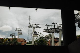 Several cable cars suspended on multiple cables traverse through a city area with some greenery and buildings. Utility poles and electrical wires are also visible against a cloudy sky. The view appears framed by an overhead structure in the foreground.