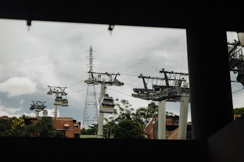 Several cable cars suspended on multiple cables traverse through a city area with some greenery and buildings. Utility poles and electrical wires are also visible against a cloudy sky. The view appears framed by an overhead structure in the foreground.