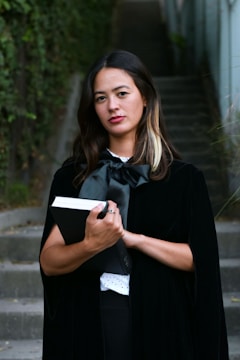 A woman stands confidently on an outdoor staircase, surrounded by greenery. She has long, dark hair with a lighter streak and is wearing a black cape and bow. She holds a thick black book, adding an academic or formal touch.