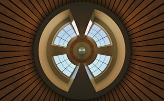 A symmetrical architectural design with concentric circles and intersecting lines, featuring a circular skylight with grid-patterned windows that allow natural light to enter. The structure exhibits a combination of geometric shapes and clean lines, with wooden slats radiating outward.