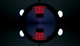 A symmetrical, kaleidoscope-like image featuring the word 'EXIT' in bold red letters repeated twice. The letters are displayed at opposite ends, surrounded by a dark circle with a gradient of blue and purple at the center. Bright circular lights appear at the four corners of the image, contributing to the geometric and symmetrical design.