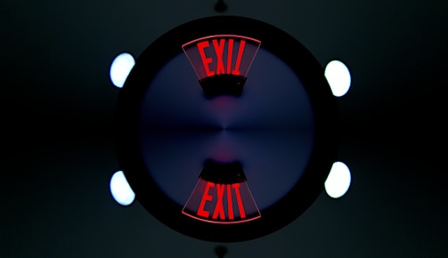 A symmetrical, kaleidoscope-like image featuring the word 'EXIT' in bold red letters repeated twice. The letters are displayed at opposite ends, surrounded by a dark circle with a gradient of blue and purple at the center. Bright circular lights appear at the four corners of the image, contributing to the geometric and symmetrical design.